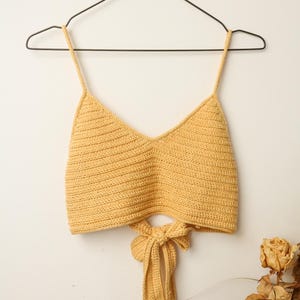 Puede incluir: Un top corto de crochet amarillo con un lazo en la espalda.