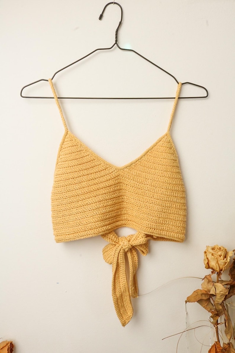 Puede incluir: Un top corto de crochet amarillo con un lazo en la espalda.