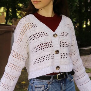 May include: A white, open-weave crochet cardigan with a V-neck and button closure. The cropped sweater features a horizontal striped pattern and is worn over a maroon top and blue jeans. The cardigan has three buttons.