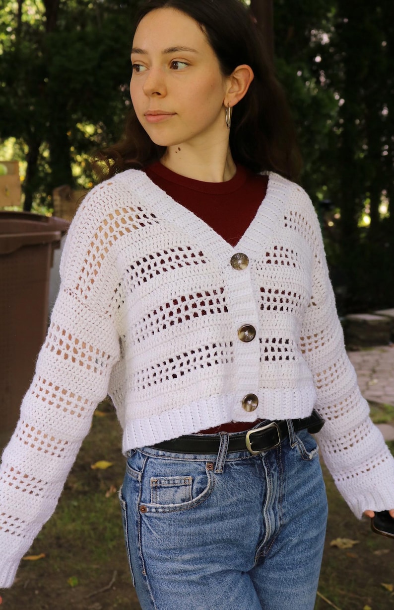 May include: A white, open-weave crochet cardigan with a V-neck and button closure. The cropped sweater features a horizontal striped pattern and is worn over a maroon top and blue jeans. The cardigan has three buttons.