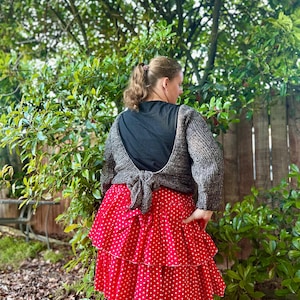 May include: A woman wearing a gray knit sweater tied in the back over a black top, paired with a red polka dot tiered skirt. The skirt has white polka dots and a white trim. The outfit is worn outdoors with a green leafy background.