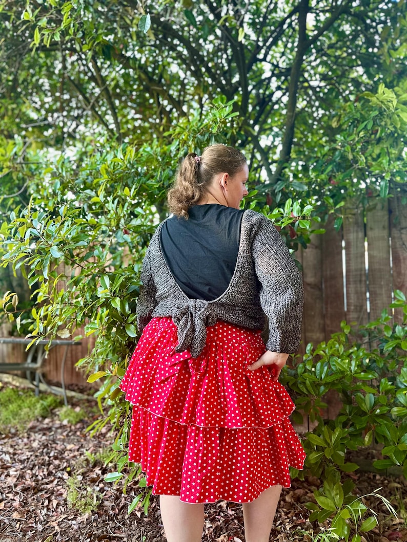 May include: A woman wearing a gray knit sweater tied in the back over a black top, paired with a red polka dot tiered skirt. The skirt has white polka dots and a white trim. The outfit is worn outdoors with a green leafy background.
