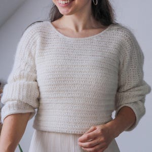 May include: Cream-colored, hand-crocheted sweater with a wide neckline and three-quarter sleeves. The sweater has a ribbed texture and a slightly cropped fit. The sleeves are cuffed. The model is wearing a cream-colored skirt.