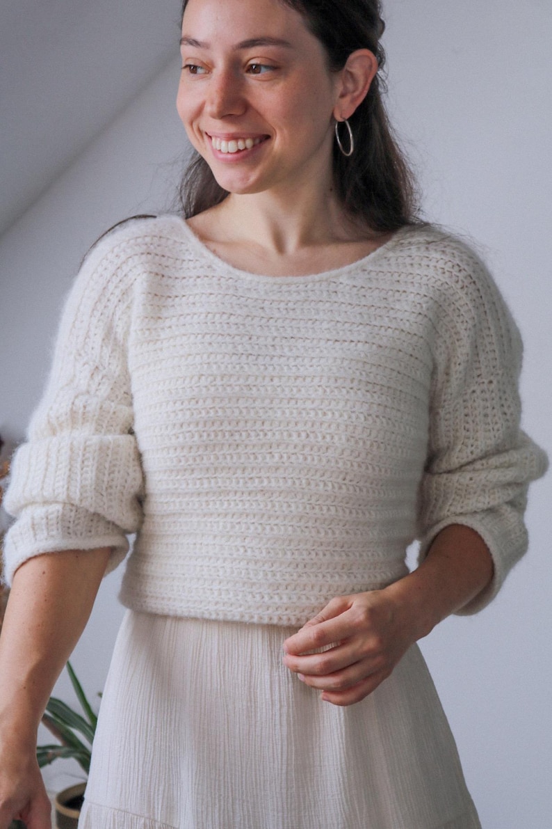 May include: Cream-colored, hand-crocheted sweater with a wide neckline and three-quarter sleeves. The sweater has a ribbed texture and a slightly cropped fit. The sleeves are cuffed. The model is wearing a cream-colored skirt.