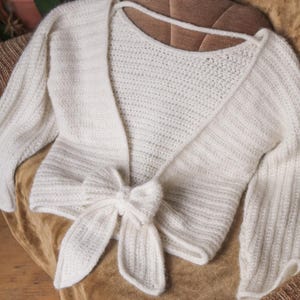 May include: A white, open-front, knitted bolero sweater with long sleeves and a tie closure at the waist. The sweater is made of a soft, textured yarn and is displayed on a brown surface.