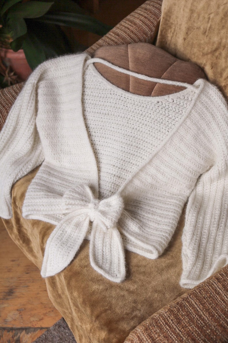 May include: A white, open-front, knitted bolero sweater with long sleeves and a tie closure at the waist. The sweater is made of a soft, textured yarn and is displayed on a brown surface.