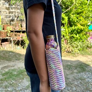 May include: A crocheted water bottle holder with a rainbow pattern and wavy stitch. The holder is made of yarn in shades of pink, purple, yellow, and green. The holder is designed to fit a standard-sized water bottle.