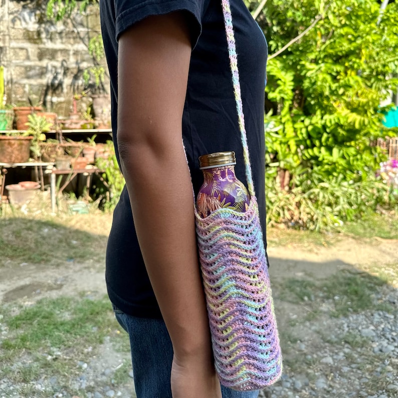 May include: A crocheted water bottle holder with a rainbow pattern and wavy stitch. The holder is made of yarn in shades of pink, purple, yellow, and green. The holder is designed to fit a standard-sized water bottle.