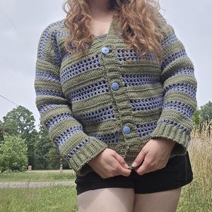 May include: A handmade crochet cardigan in shades of olive green and lavender. The open-weave design features horizontal stripes and is fastened with three light blue buttons. The sleeves and cuffs are solid olive green.