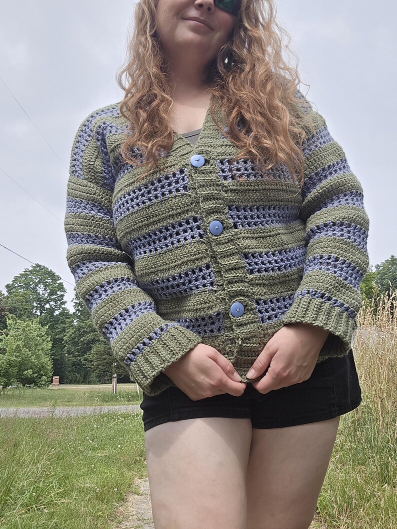 May include: A handmade crochet cardigan in shades of olive green and lavender. The open-weave design features horizontal stripes and is fastened with three light blue buttons. The sleeves and cuffs are solid olive green.