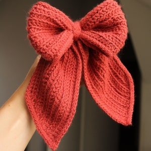 May include: A large, red crocheted bow with two long tails. The bow is tied in a knot in the centre and the tails are pointed at the ends.