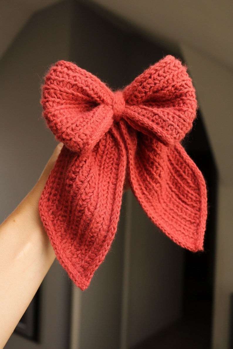 May include: A large, red crocheted bow with two long tails. The bow is tied in a knot in the centre and the tails are pointed at the ends.