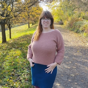 May include: A woman wearing a dusty rose crochet sweater with a boat neck and three-quarter sleeves, paired with a dark teal skirt and purple tights. The background features a path and trees with yellow leaves.
