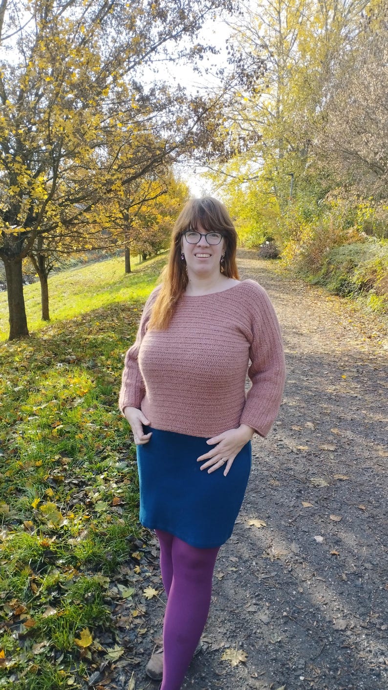 May include: A woman wearing a dusty rose crochet sweater with a boat neck and three-quarter sleeves, paired with a dark teal skirt and purple tights. The background features a path and trees with yellow leaves.