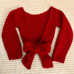 May include: A red crochet shrug with long sleeves and a large bow tie closure. The handmade bolero-style sweater is a vibrant shade of red, perfect for layering. The open front design adds a touch of elegance to the garment.