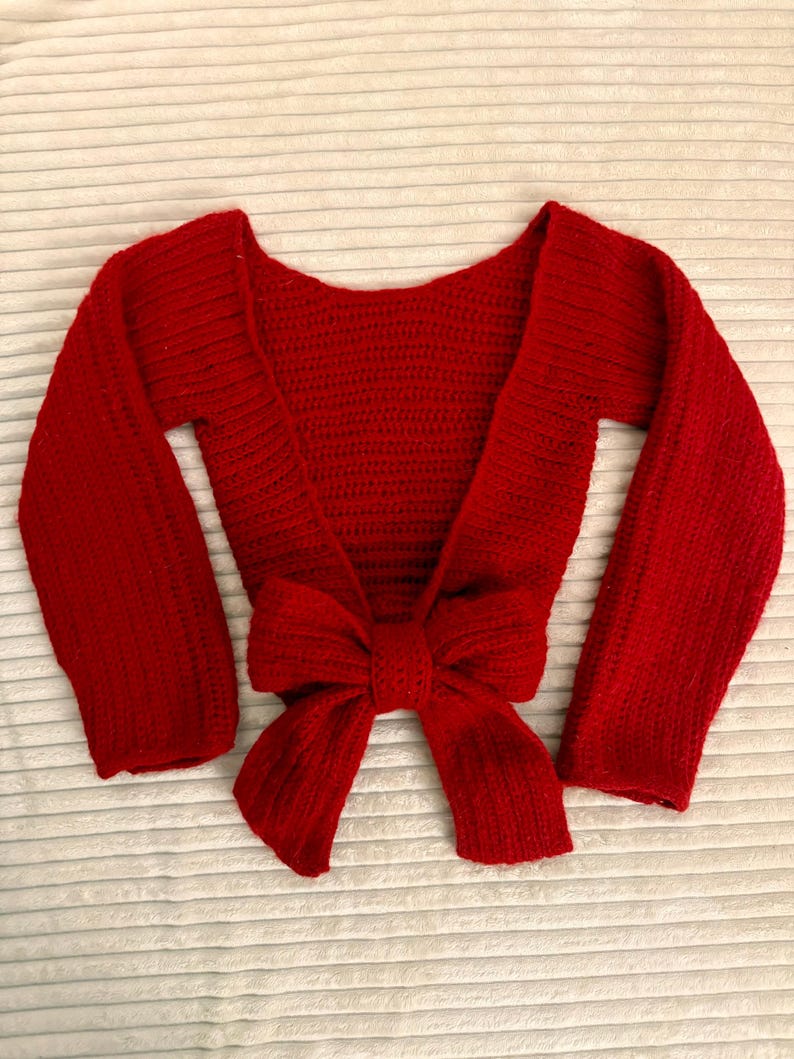 May include: A red crochet shrug with long sleeves and a large bow tie closure. The handmade bolero-style sweater is a vibrant shade of red, perfect for layering. The open front design adds a touch of elegance to the garment.