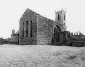 May include: Black and white image of an old stone church ruin. The building has a tall tower and arched doorways. The structure is surrounded by a grassy field and a stone wall. The sky is overcast.
