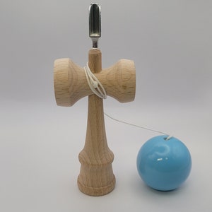 May include: A wooden kendama toy with a light blue ball attached to a string. The kendama has a silver metal spike and a wooden base.