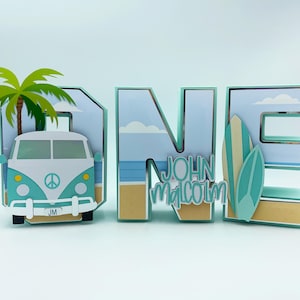 Op de afbeelding: Een 'ONE'-bord met strandthema. Voorzien van een teal en witte bus, palmboom en surfplanken. De letter 'N' heeft de naam 'John Malcolm'. Ideaal voor een eerste verjaardag.