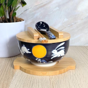 May include: A blue and white ceramic bowl with a moon and rabbit design, a wooden lid, chopsticks, and a spoon. The bowl is sitting on a wooden stand.