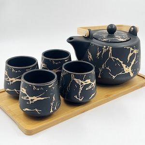 May include: Black and gold tea set with a teapot and four cups on a wooden tray. The teapot and cups have a marble pattern with gold accents.