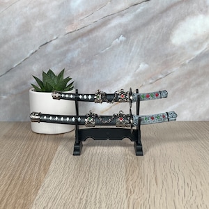 May include: Two black and silver decorative swords with red and green accents are displayed on a black stand. The swords are on a wooden surface.