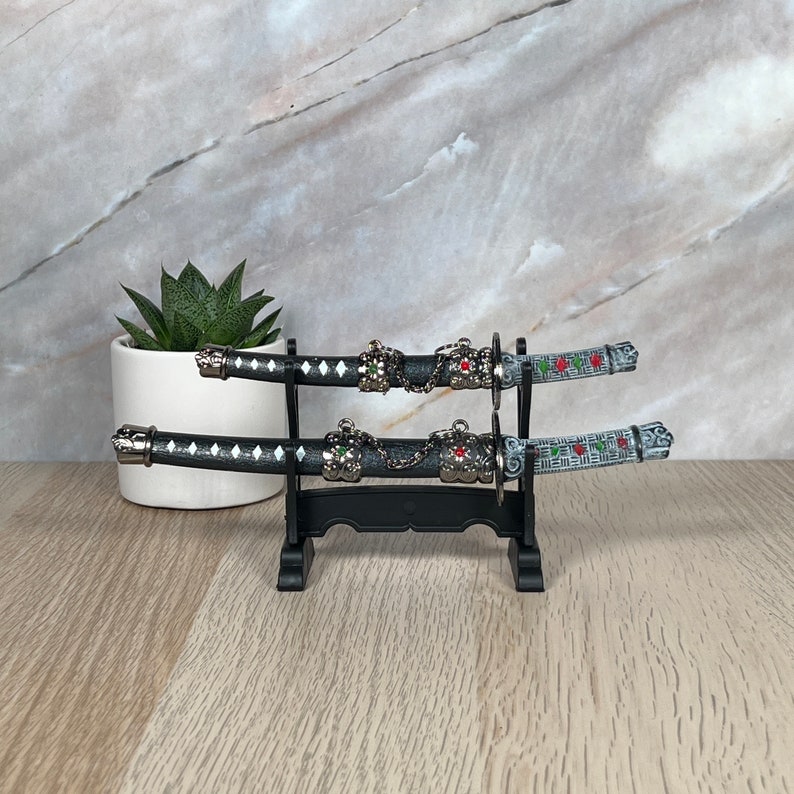 May include: Two black and silver decorative swords with red and green accents are displayed on a black stand. The swords are on a wooden surface.