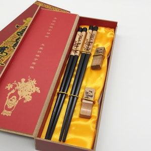 May include: A set of black chopsticks with decorative wooden tips in a red and gold box. The box has Chinese characters on it and a yellow satin lining.