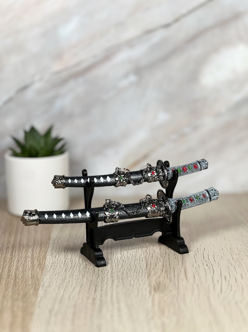 May include: Two black and silver miniature samurai swords with red and green accents are displayed on a black stand. The swords have a white and black patterned design.