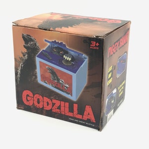 May include: Godzilla-themed piggy bank in a box. The box is brown with a Godzilla graphic and the word "GODZILLA" in red. The piggy bank has a blue and white design with a Godzilla graphic. The box also says "Vivid and Great in Style."