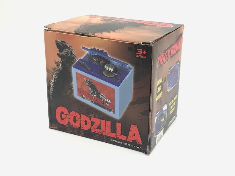 May include: Godzilla-themed piggy bank in a box. The box is brown with a Godzilla graphic and the word "GODZILLA" in red. The piggy bank has a blue and white design with a Godzilla graphic. The box also says "Vivid and Great in Style."