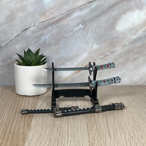 May include: Three decorative swords with black and silver handles and red and green accents are displayed on a black stand. The swords are on a wooden surface.