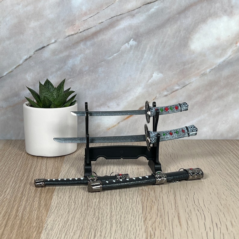 May include: Three decorative swords with black and silver handles and red and green accents are displayed on a black stand. The swords are on a wooden surface.