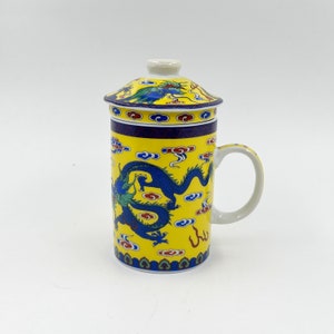 May include: A yellow ceramic teacup with a lid, featuring a blue dragon design with red and green accents. The cup has a handle and a blue and white border.