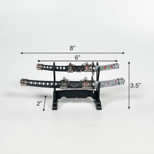 May include: Two black and silver decorative swords with green and red accents on a black stand. The swords are 6 inches long and the stand is 2 inches tall. The swords are displayed on a white background.