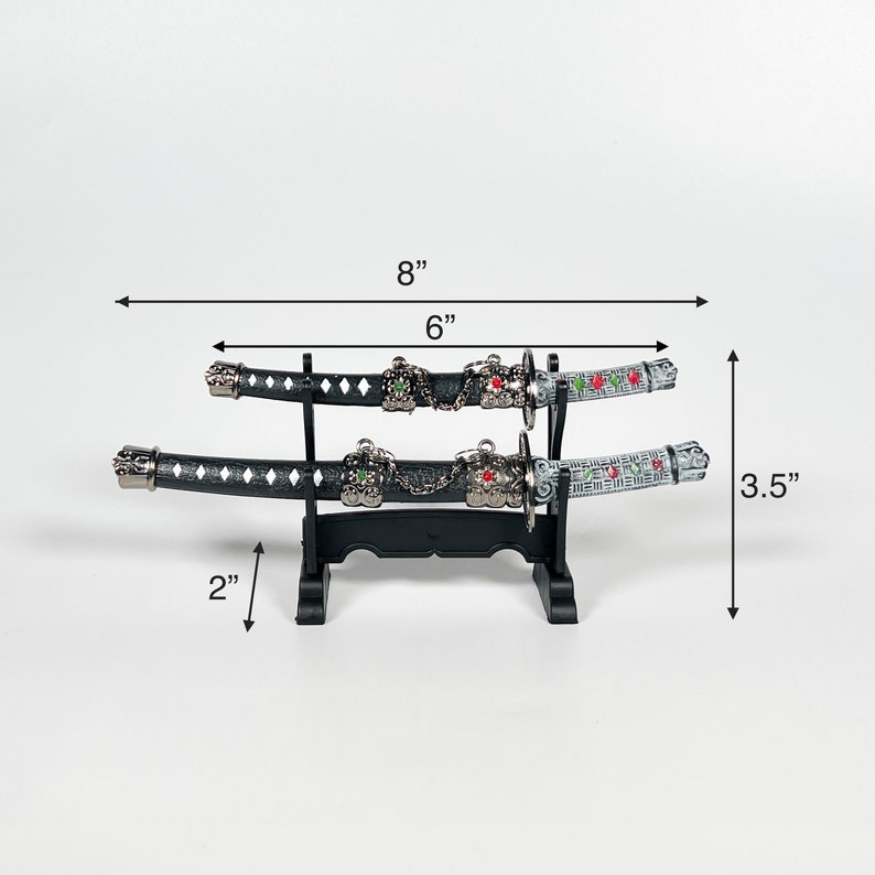 May include: Two black and silver decorative swords with green and red accents on a black stand. The swords are 6 inches long and the stand is 2 inches tall. The swords are displayed on a white background.