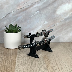 May include: Two black and silver decorative swords with red and green accents are displayed on a black stand. The swords are on a wooden surface.