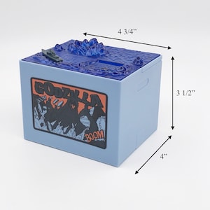 May include: A light blue Godzilla-themed bank, 4" x 4 3/4" x 3 1/2", with a detailed ocean scene on top and a Godzilla graphic on the side. The bank features a slot for inserting money and a small boat.