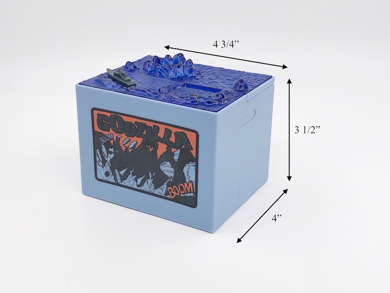 May include: A light blue Godzilla-themed bank, 4" x 4 3/4" x 3 1/2", with a detailed ocean scene on top and a Godzilla graphic on the side. The bank features a slot for inserting money and a small boat.