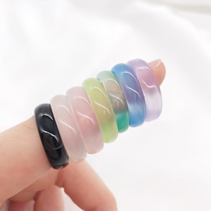 Pode incluir: Um conjunto de sete anéis coloridos feitos de pedra lisa e polida. Os anéis estão empilhados uns sobre os outros, com o anel preto na parte inferior e o anel roxo claro na parte superior.