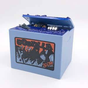 May include: A light blue Godzilla-themed box with a hinged lid. The lid is open, revealing a monster emerging from blue water. The box features a Godzilla graphic and the word "GODZILLA" in orange and black.