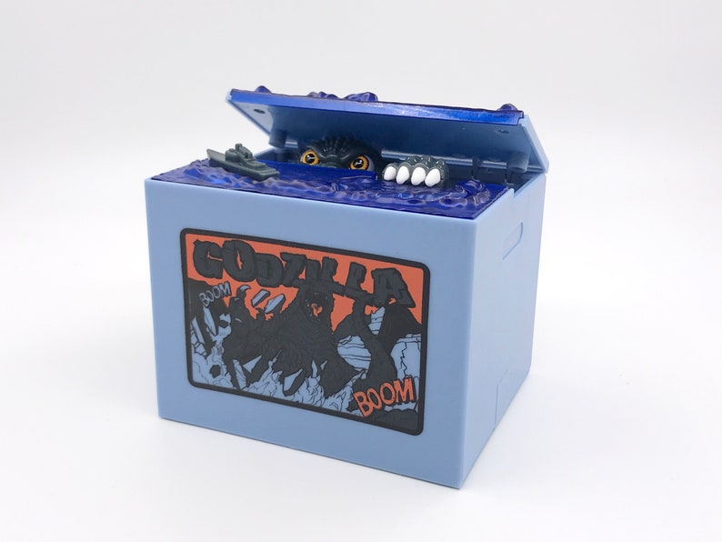 May include: A light blue Godzilla-themed box with a hinged lid. The lid is open, revealing a monster emerging from blue water. The box features a Godzilla graphic and the word "GODZILLA" in orange and black.