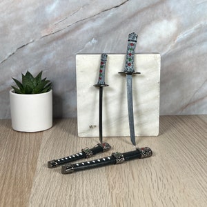 May include: Two miniature swords with black scabbards and silver accents. The swords have silver and black handles with red and green accents. The swords are displayed on a white marble surface.