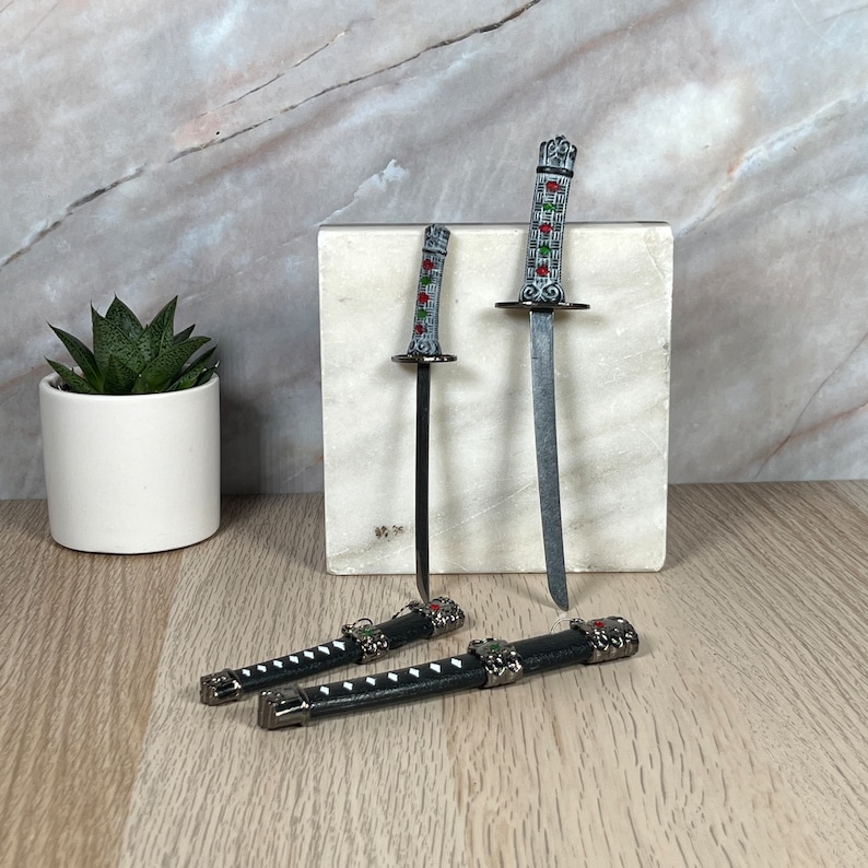 May include: Two miniature swords with black scabbards and silver accents. The swords have silver and black handles with red and green accents. The swords are displayed on a white marble surface.