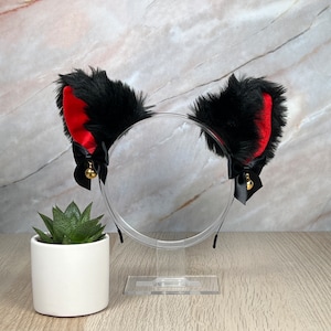 May include: Black cat ears headband with red velvet trim and gold bells. The headband is on a clear stand.