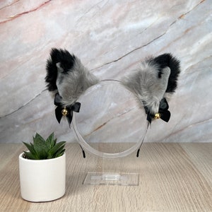 May include: Gray and black furry cat ears headband with black bows and gold bells. The headband is on a clear plastic stand.