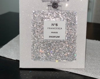 Puede incluir: Un lienzo blanco con un fondo de purpurina plateada brillante. Una botella de vidrio transparente con una tapa plateada está sobre el lienzo. La botella tiene una etiqueta blanca con el texto "N°5 FRANCESCA PARIS PARFUM".