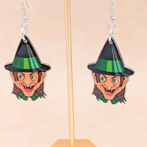 May include: A pair of Halloween-themed earrings featuring a cartoon witch face. The earrings have a black witch hat with a green band, green eyes, and a smiling face. The earrings are hanging from silver hooks.