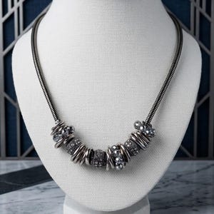 May include: A silver-toned necklace with a flexible chain and decorative beads. The necklace features a series of silver rings and sparkling, dark gray beads. The necklace is displayed on a white mannequin.