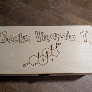 Puede incluir: Una caja de madera de color claro con las palabras "Becks Vitamin T" grabadas en la parte superior. Debajo del texto hay un diagrama molecular. La caja tiene un pequeño cierre de metal.